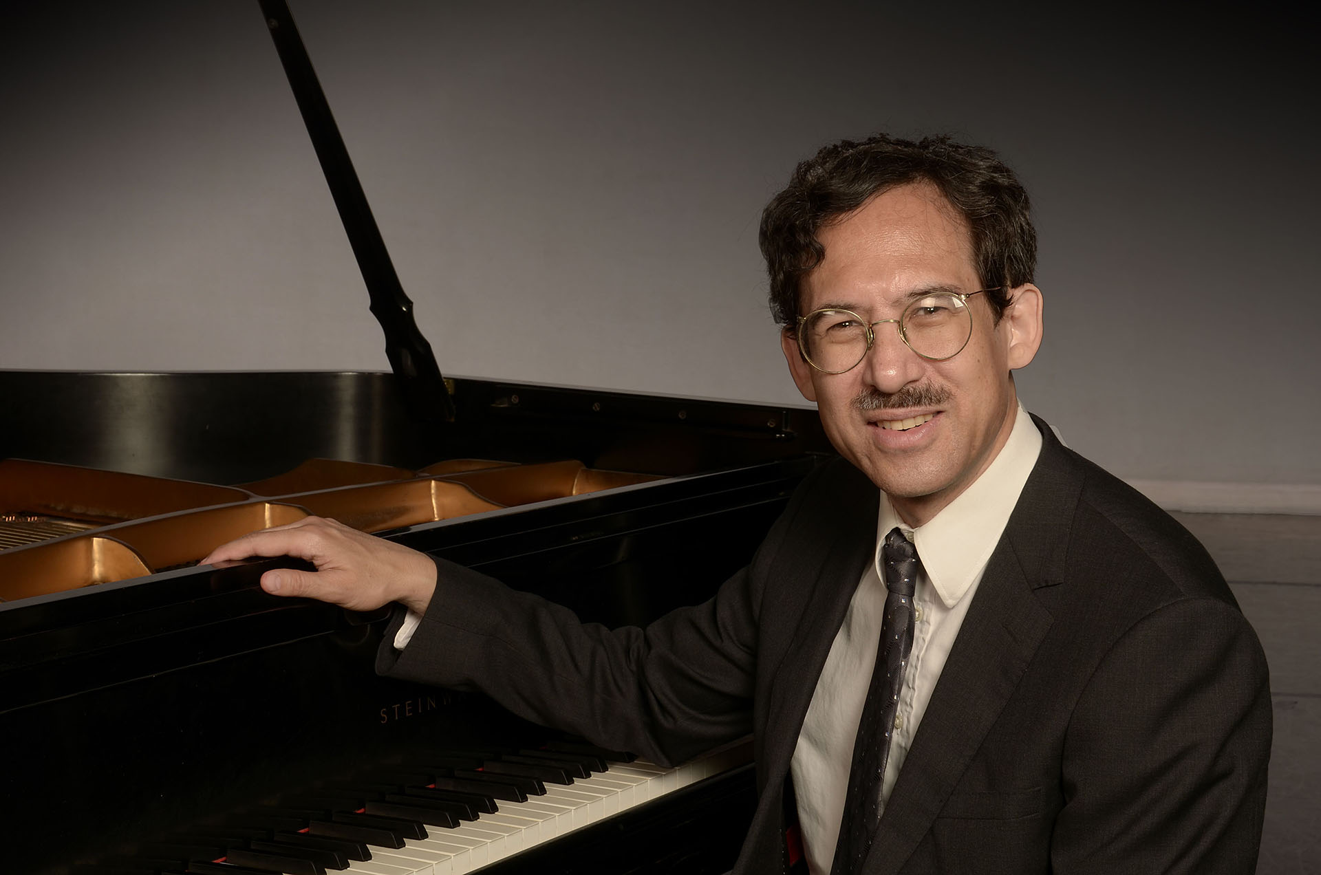 Photo of Michael Arnowitt in dark gray suit seated resting arm on grand piano
